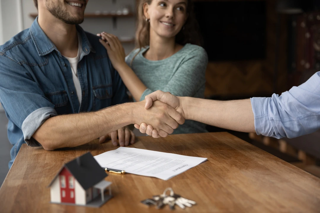 A close-up of two hands shaking over a table with a contract and a miniature house, symbolizing a real estate agreement. A close-up of two hands shaking over a table with a contract and a miniature house, symbolizing a real estate agreement.