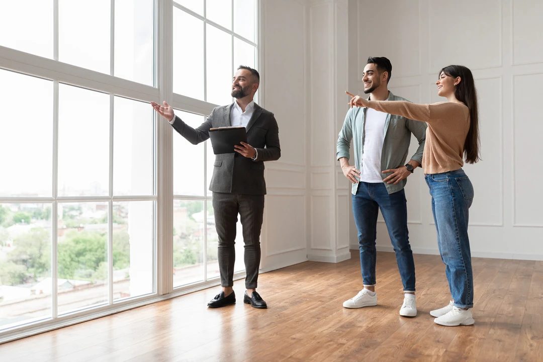A real estate agent gestures towards large windows while showing a property to a couple in a spacious, well-lit room. A real estate agent gestures towards large windows while showing a property to a couple in a spacious, well-lit room.